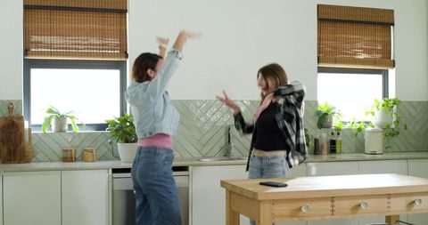 Friends dancing and laughing in sunlit modern kitchen wearing jeans and casual tops