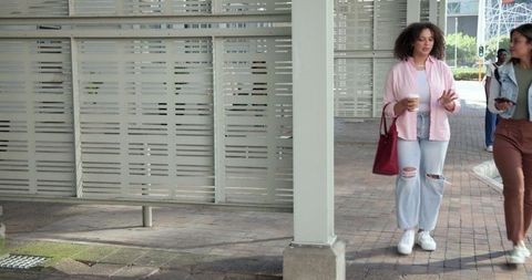 Diverse female friends walking with coffee and red tote on urban plaza walkway chatting
