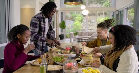 Friends Carving Roast Chicken and Sharing Salad Around Sunlit Modern Dining Table