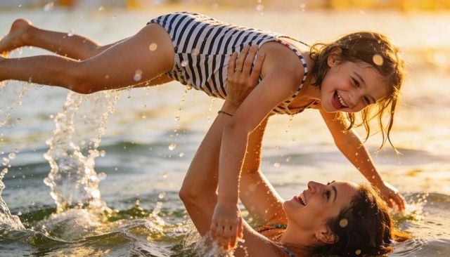 Mother lifting laughing daughter in sunlit sea at sunset, playful summer family moment