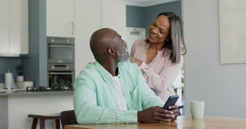 Mature couple sharing smartphone moment in modern sunlit kitchen, smiling and connecting