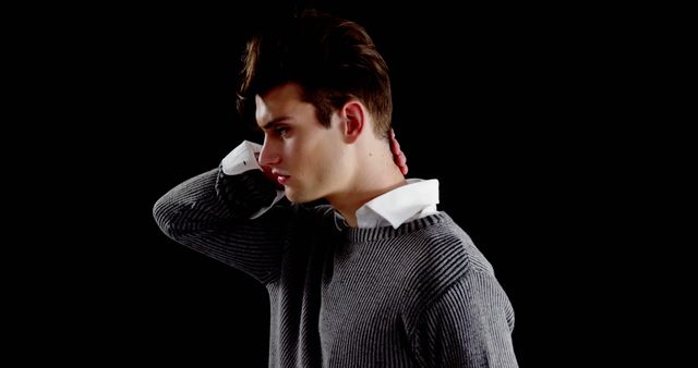 Young man posing in dark lighting wearing a gray sweater over a white shirt and touching his neck. Suitable for use in fashion promotions, model portfolios, and styling guides.