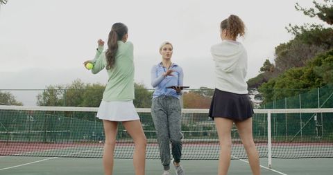 Female tennis coach instructing two players on outdoor court near net with tablet