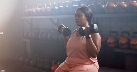 African american woman performing seated dumbbell shoulder press on bench with kettlebells