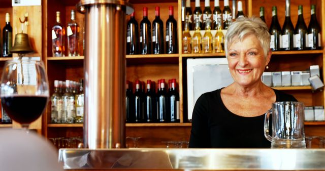 Elderly woman in black shirt smiling warmly while standing behind bar counter in winery. Background filled with wine bottles on wooden shelves, highlighting cozy and welcoming atmosphere. Perfect for themes related to hospitality, customer service, senior employment, and small business management.