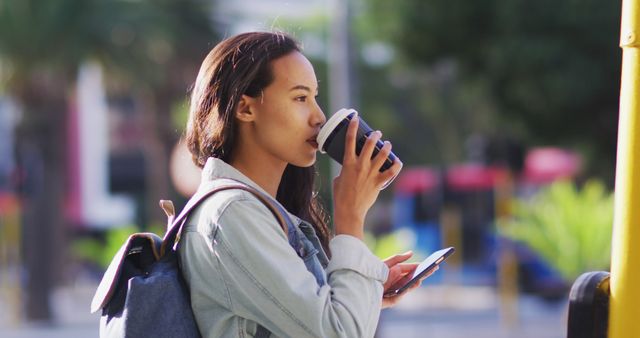 Young woman holding a coffee cup and smartphone, wearing casual clothes with a backpack in an urban outdoor environment. Perfect for themes related to modern lifestyle, city life, technology use, or casual street scenes.