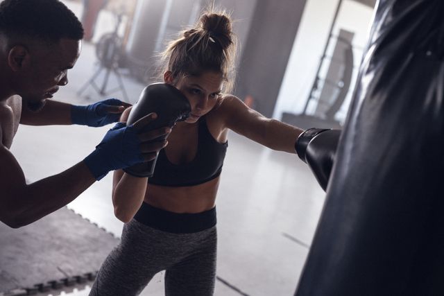 Multiracial male coach guiding and training female boxer punching boxing bag in health club. Glove, guidance, togetherness, unaltered, boxing, sport, training, strength and fitness concept.