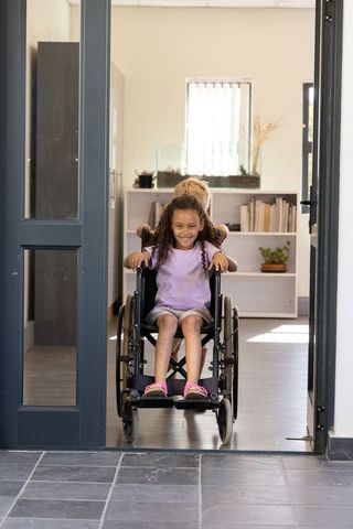 Caregiver pushing smiling african american girl in wheelchair through bright home doorway