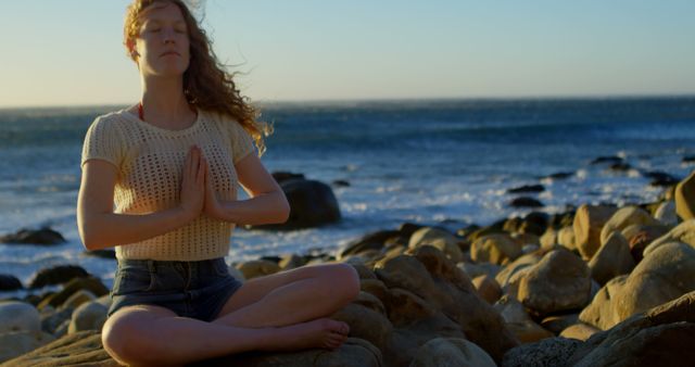 Young Caucasian woman meditates on a rocky beach at sunset. She practices mindfulness in a serene outdoor setting by the ocean.