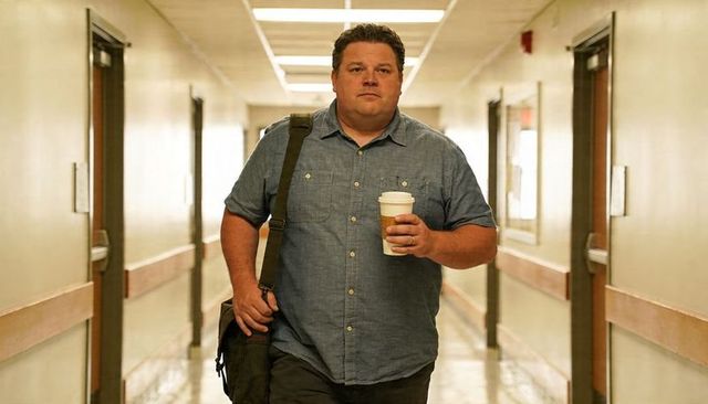 Man walking hospital corridor carrying coffee and shoulder bag in chambray shirt