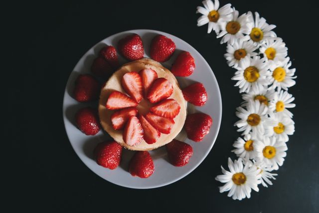 Perfect for use in food blogs, recipe books, bakery advertisements, and culinary artwork, this vibrant image features a strawberry shortcake garnished with sliced and whole strawberries on a plate, accented with a bouquet of white daisies arranged on a dark background. Ideal for themes involving desserts, freshness, and gourmet presentations.