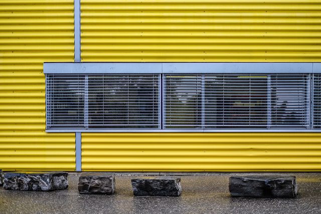 Yellow industrial building with metal louver shutters on window creating modern urban design. Concrete blocks line base against yellow corrugated wall. Ideal for illustrating urban development, industrial architecture, or construction themes.