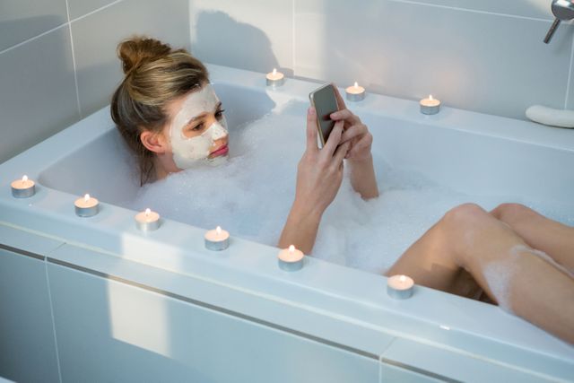 Woman enjoying a bubble bath while using a mobile phone, surrounded by lit candles. Ideal for concepts related to self-care, relaxation, home spa treatments, and wellness routines. Perfect for promoting beauty products, spa services, and lifestyle blogs.