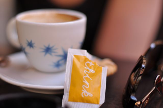 Close-up of a coffee cup on a table with an artistic design, accompanied by a sugar packet labeled 'think'. Suitable for depicting morning routines, cozy cafe settings, or moments of contemplation and inspiration. Ideal for use in food and beverage promotions, blogs about coffee culture, or advertising for cafes and restaurants.