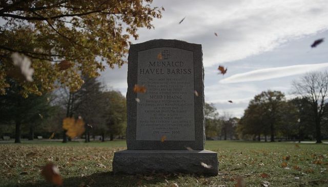 Falling autumn leaves swirling around granite headstone with carved cross in park cemetery