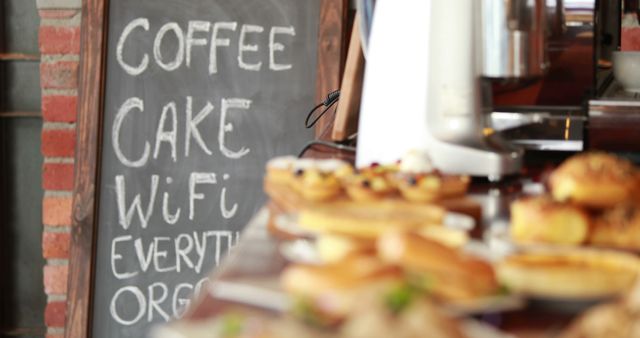A variety of pastries are displayed in the foreground at a cafe, with a chalkboard menu in the background listing coffee, cake, and WiFi availability, with copy space. The focus on the food suggests a cozy and inviting atmosphere for customers looking for refreshments and connectivity.
