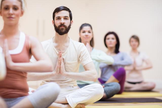 Group of adults practicing yoga in a fitness studio, sitting in lotus position with hands in prayer pose. Ideal for promoting wellness, mindfulness, and healthy lifestyle activities. Suitable for use in fitness, health, and wellness-related content.