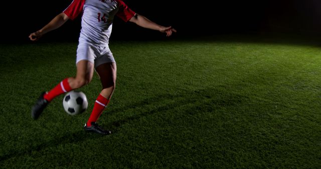Soccer player is kicking the ball on a grassy field during a night game. The intense moment is captured mid-action, highlighting the dynamics and skill involved. The athlete is in a competitive stance, making it ideal for sports articles, advertisements for soccer gear, or illustrating athleticism in visual presentations.