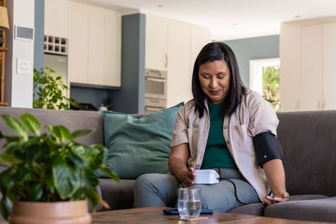 Mature woman checking blood pressure at home using digital monitor on living room sofa