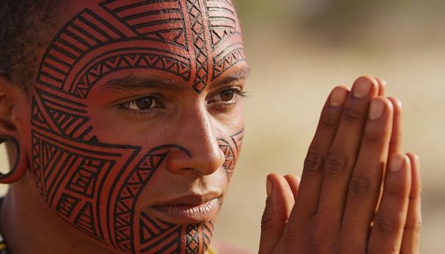 Sunlit contemplative man with geometric facial tattoos pressing hands together