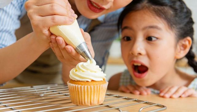 Parent guiding piping bag while child watching cupcake frosting and decorating at home