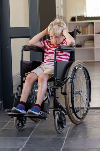 Upset blonde boy sitting in wheelchair covering ears in minimalist home hallway