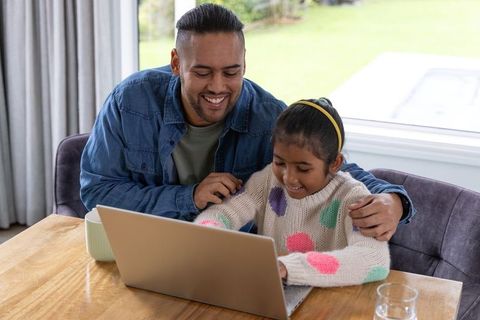 Father guiding daughter on laptop, smiling and bonding during cozy family home learning moment