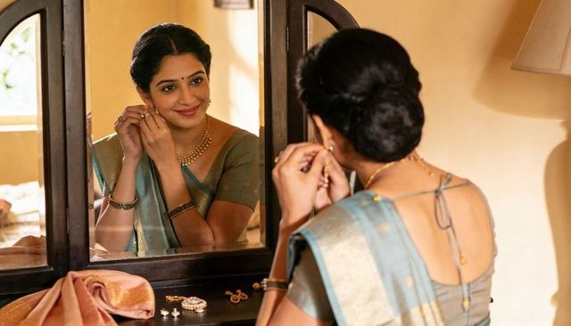 South asian woman putting on gold earring in teal silk saree at wooden vanity mirror