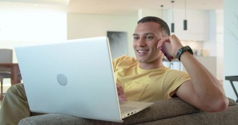 Young man relaxing on couch using laptop and talking on smartphone in modern sunlit apartment