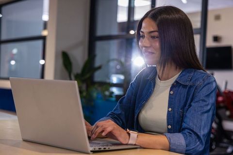 Asian woman typing on laptop in modern coworking space wearing smartwatch and denim shirt