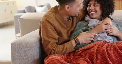 African american couple cuddling on gray sofa under cozy rust-red blanket, sharing warm smiles