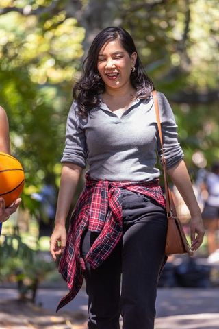 Smiling woman walking park with plaid tied at waist carrying tan bag, companion holding basketball