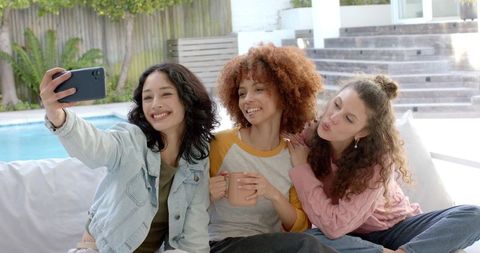 Three Friends Taking Selfie on Poolside Sofa Drinking Coffee and Laughing Outdoors