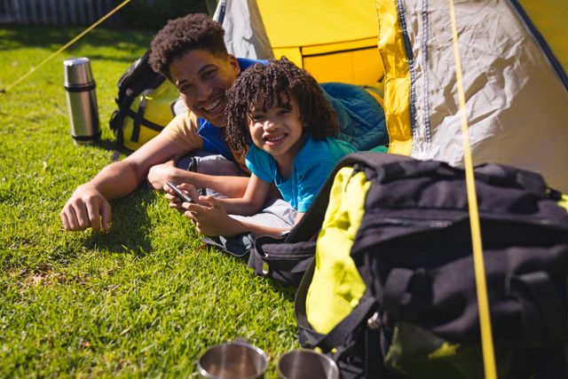 Hispanic father and son enjoying a sunny day camping in a backyard tent. Perfect for illustrating family bonding, outdoor activities, and summer leisure. Ideal for use in lifestyle blogs, family-oriented advertisements, and outdoor adventure promotions.
