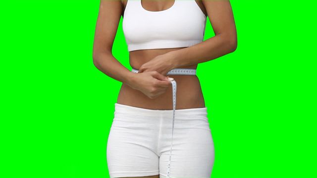 Woman in white sportswear measuring her waistline with a tape measure against green chroma key background. Useful for health and fitness promotion, weight loss programs, wellness articles, and body positivity campaigns.