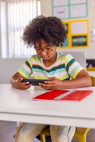 African american student playing on handheld device at desk with red notebook