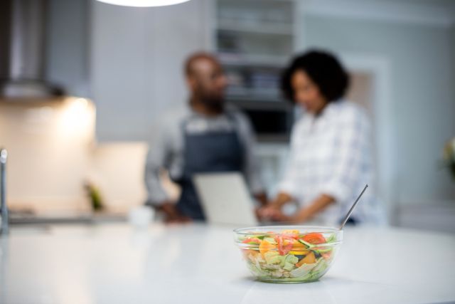 Bowl of fresh salad on kitchen counter with couple in background. Ideal for promoting healthy eating, lifestyle blogs, cooking websites, and domestic life themes.