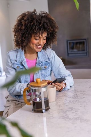 Smiling african american woman texting at marble kitchen island with french press coffee