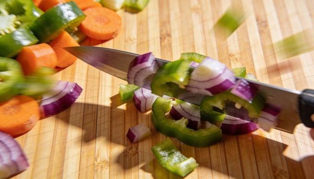Sharp chef knife slicing red onion, green bell pepper and carrot on bamboo cutting board