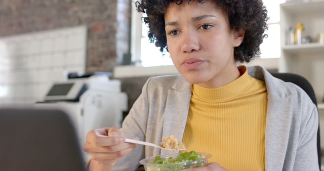 Young African American businesswoman wearing a yellow sweater intently watching something on a computer screen while eating a salad in an office environment. This is useful for illustrating modern work culture, healthy eating habits, and the multitasking nature of professional life.