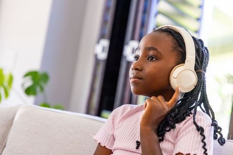 African girl listening on sofa with cream headphones, concentrating in bright living room