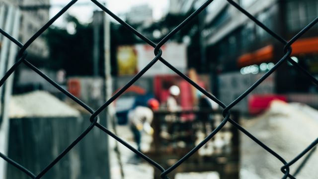 Construction workers are seen behind a chain-link fence, focusing on the fence while the background is blurred. This evokes themes of security, boundaries, and urban development. Ideal for topics related to construction industry, urban growth, and security.