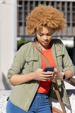 Young african american woman leaning on urban railing checking smartphone in sunlit city