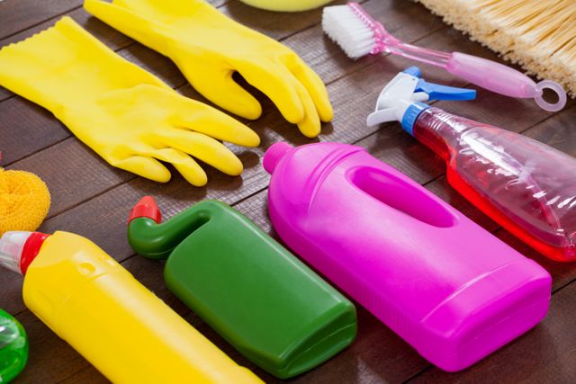 This image shows an assortment of cleaning supplies neatly arranged on a wooden floor. Items include rubber gloves, spray bottles, detergent bottles, and a scrub brush, all in vibrant colors. Ideal for use in articles, blogs, and advertisements related to home cleaning, hygiene, and household maintenance. This image portrays cleanliness and the tools needed to achieve it.