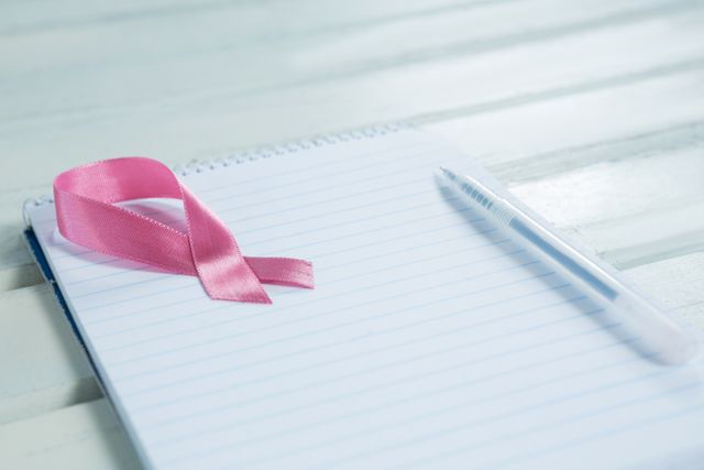 Pink Breast Cancer Awareness ribbon placed on a spiral notepad with a pen on a wooden table. Ideal for use in health campaigns, educational materials, charity events, and awareness promotions.