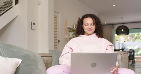 African american woman in pink hoodie using laptop on sofa for remote work and home study
