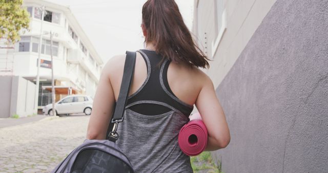 A woman carrying a yoga mat and gym bag walking in an urban area. She is wearing sportswear, likely heading to a workout or yoga session. This can be used to illustrate themes of fitness, healthy lifestyle, exercise routines, and urban fitness locations.
