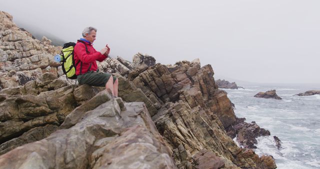 Mature man hiking on a rocky coastline capturing the scenic view with his smartphone. He is wearing a red jacket and a green backpack, sitting on large rock formations near the ocean. Ideal for use in travel and tourism promotions, adventure and outdoor lifestyle blogs, and articles related to nature exploration and senior outdoor activities.