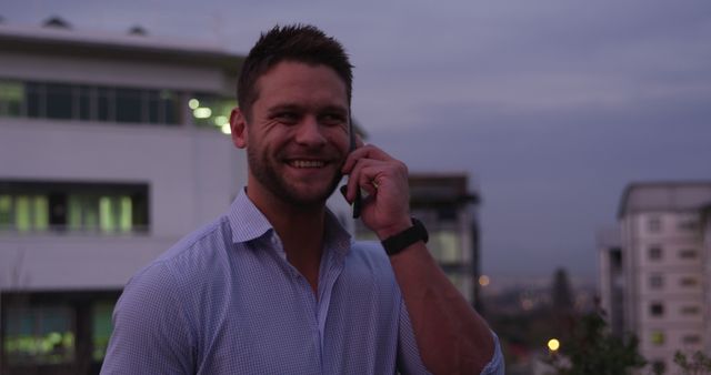 Man enjoying a phone conversation outside office buildings during the evening. Suitable for business communication themes, technology use, work-life balance, and urban lifestyle applications.