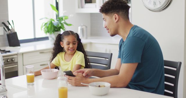 Happy biracial father and daughter eating breakfast and using smartphone in kitchen. domestic lifestyle, spending free time at home.